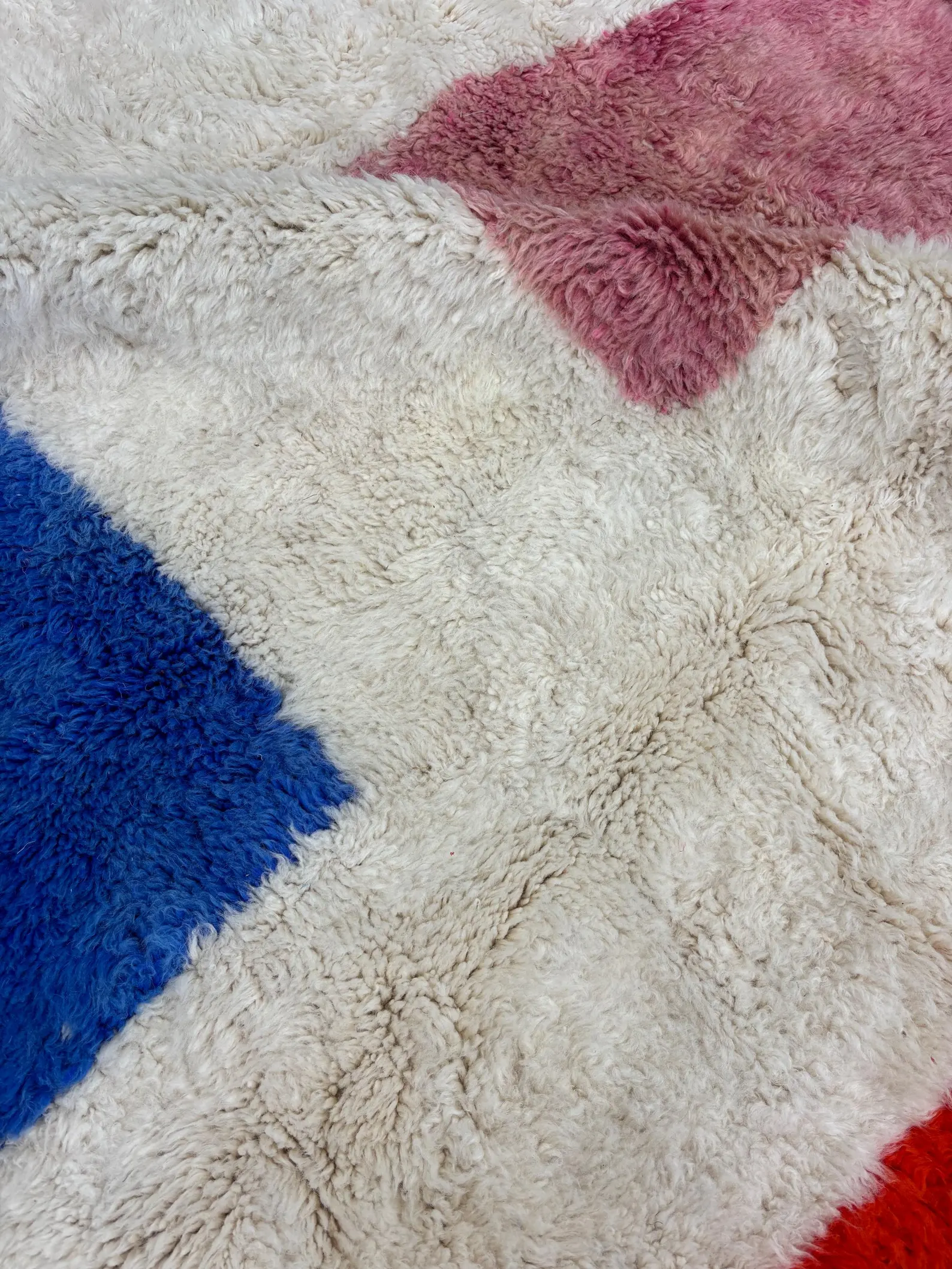 Close-up of a fluffy shag rug with blue, pink, and red color patches on a beige base, showing soft textured fibers.